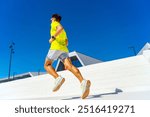 A runner in yellow gear is captured from behind, ascending steps under a vibrant, clear blue sky.

