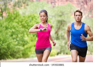 Runners - Mixed Multicultural Couple Training Outside In Nature. Fitness Asian Young Smiling Model Checking Time Or Pulse On Heart Rate Monitor Watch And Male Model Jogging.