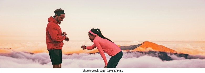Runners tired breathing after training looking at their wearable tech sports watch checking heart rate health data. Two athletes couple running together in outdoors mountain banner panorama.