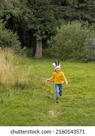 Running Boy Is Playing American Indian On Field. Kid Has Handmade Headdress Made Of Feathers And Bow With Arrows. Costume Role Play. Outdoor Leisure Activity. Fall Season.