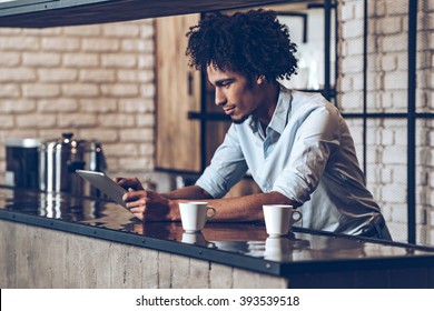 Running business easier with technologies. Side view of young African man using his digital tablet while leaning at bar counter with two coffee cups - Powered by Shutterstock