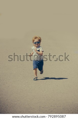 Running child among lot of sand. Joyful cute kid in desert, dynamic boy in T-shirt, shorts, sandals and blue sunglasses isolated sienna background. Running in desert, sport on beach, healthy lifestyle