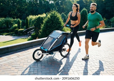 Running couple with child in jogging stroller in public park at sunrise. Morning run for a whole family