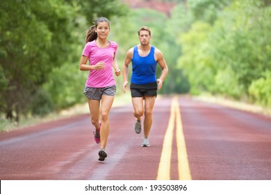 Running Couple Training Outdoors On Road In Wood For Marathon. Mixed Race Pretty Young Female And Fit Caucasian Handsome Man In Their Twenties. Lifestyle Full Figure