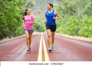 Running - Exercising Couple Jogging And Talking Outside On Road Beautiful Nature Landscape. Runners Training Together For Marathon Run. Asian Female Sports Woman And Male Fitness Man In Full Body.