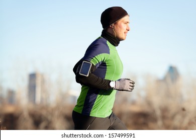 Running Man Jogging In Autumn Listening To Music On Smart Phone. Runner Training In Warm Outfit On Cold Day. Fit Male Fitness Athlete Model Training Outdoor In Fall. Full Body Length Of Jogger.