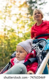 Running Mother With Contemplative Child In Stroller Enjoying Motherhood At Autumn Sunset And Forest Landscape. Jogging Or Power Walking Woman With Pram In Woods.