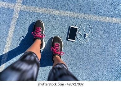 Running Shoes Girl Feet Selfie On Blue Track Lane Getting Ready To Run With Smartphone And Earbuds To Listen To Mobile Phone Music App. POV Of Runner Woman Athlete And Her Fit Legs In Activewear.