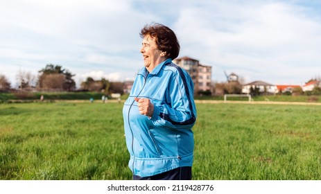 Running And Sports Activity. An Elderly Smiling Woman In Sports Clothes Runs Through A Green Field. Side View. The Concept Of The International Day Of Older Persons.