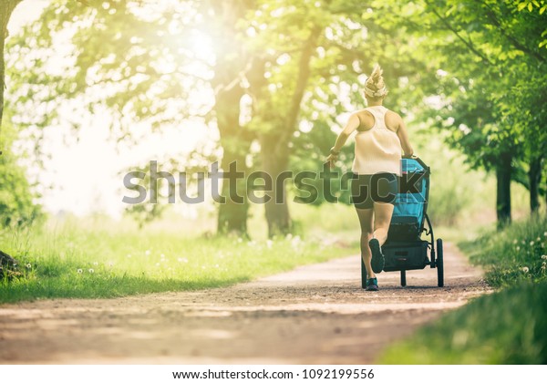 Running woman with baby stroller enjoying summer\
day in park. Jogging or power walking supermom, active family with\
baby jogger.