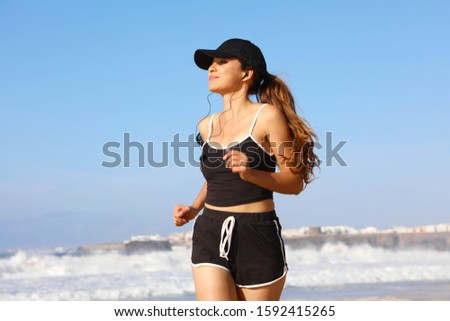 Running woman with black cap listening to music with earphones on smart phone in summer beach