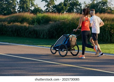 Running Young Family With Baby Jogging Stroller In Motion Enjoying Workout And Summer Day In City Park. Back View