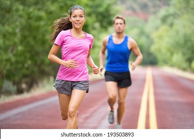 Running Young Mixed Race Couple Training For Marathon Outside On Road. Happy Smiling Beautiful Mixed Race Female Model In Front - Handsome Caucasian Male Model Blurred In Background, Running Focused