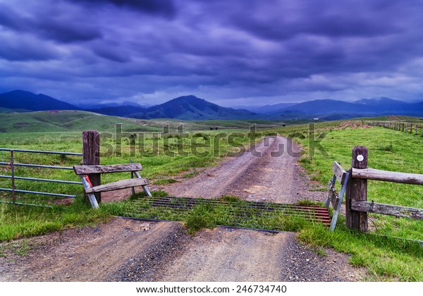 Rural agricultural property\
dividing fence, grid and gate on unsealed road through property in\
stormy valley with green grazing grass surrounded by hilly\
mountains