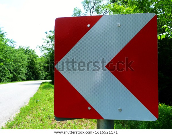 rural asphalt\
road. sharp curve ahead. road side metal warning  sign metal square\
arrow in red and white, lush green foliage and trees in the \
background. road safety\
concept.