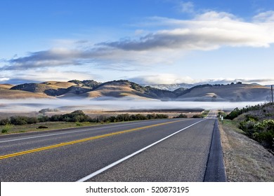 A Rural California Road, In A Valley, Shrouded, In A Layer Of Fog At Sunrise, With Blue Sky, White Clouds & Gentle Rolling Hills In The Background. Photographed On The California Central Coast.