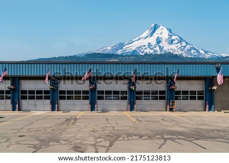 Rural firehouse station housing multiple fire trucks in individual garage door bays with American flags and snow covered mountain the background.
