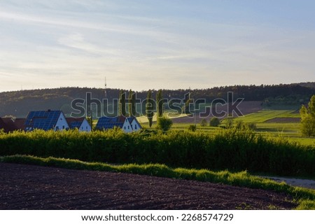 Rural houses with solar panels on the roofs. In the foreground is a plowed agricultural field. Fields and forests in the background. Rural landscape