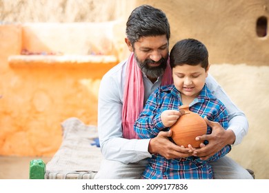 Rural Indian Father And Son Putting Coin Into Clay Money Box. Happy Beard Man And Boy Holding Traditional Piggy Bank Or Gullak, Dad Teaching Kid To Save Money, Investment And Banking Concept.