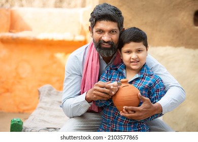 Rural Indian father and son putting coin into clay money box. Happy beard man and boy holding traditional piggy bank or Gullak, dad teaching kid to save money, Investment and banking concept.