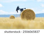 Rural landscape in prairies with big round yellow bales of fescue straw and black oil pump jack sin the field under blue cloudy sky.