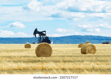 Rural landscape in prairies with big round yellow bales of fescue straw and black oil pump jack sin the field under blue cloudy sky. - Powered by Shutterstock