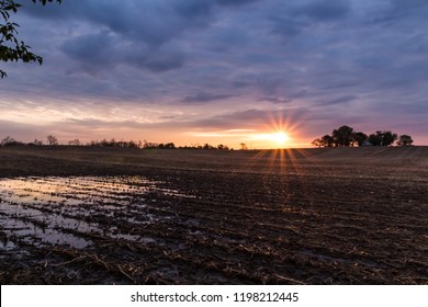 Rural Landscape Scene Of Sunrise Sunset Over A Farm Field After Harvest. Sun Breaks Through Clouds Creating Reflection In Water Standing In The Field. Concepts Of Family Farm, Winter, After Harvest