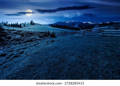rural landscape in spring at night. haystack near wooden fence on green meadow in full moon light. spruce forest on rolling hills. mountain with snow tops. finance investment or abstract concept