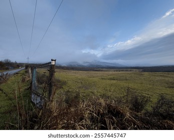 Rural Landscape with Weathered Fence, Open Field, and Distant Mountain Covered in Mist Under Cloudy Sky. Pásztó, Muzsla - Powered by Shutterstock