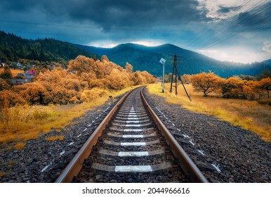 Rural railroad in mountains in overcast day in autumn. Old railway station in village at sunset. Industrial landscape with railway platform, orange trees and grass, dramatic cloudy sky, buildings