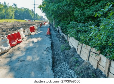 A rural road undergoing construction bordered by a dense green tree line. Red and white plastic safety barriers line the paved roadway while a trench filled