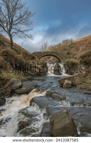 A rural winter landscape scene of the waterfall and packhorse stone bridge at Three Shires Head in the Peak District National Park.