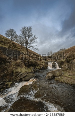 A rural winter landscape scene of the waterfall and packhorse stone bridge at Three Shires Head in the Peak District National Park.