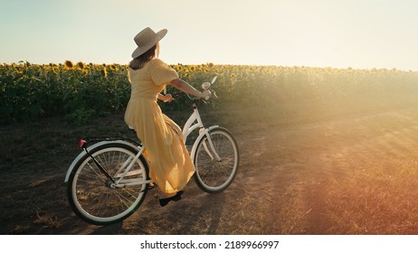 Rural Woman In Timeless Dress Riding Retro Styled White Bicycle On Country Road Alone Near Sunflowers Field. Vintage Fashion, Amazing Adventure, Countryside Activity, Healthy Lifestyle.