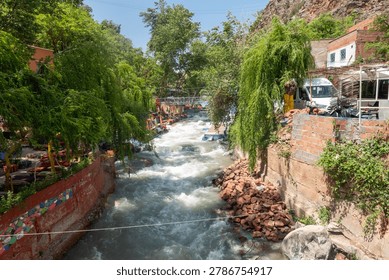 A rushing mountain river flows through the scenic Ourika Valley in Morocco, lined with lush green trees, local cafes, and rustic buildings under a bright sky.