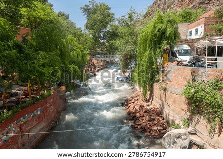 A rushing mountain river flows through the scenic Ourika Valley in Morocco, lined with lush green trees, local cafes, and rustic buildings under a bright sky.