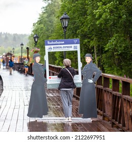 Ruskeala Park, Republic Of Karelia, Russia - July, 2021: A Woman Stands At A Selfie Frame Made In The Form Of An Instagram Frame With Two Charming Train Conductors Dressed In The Old Era