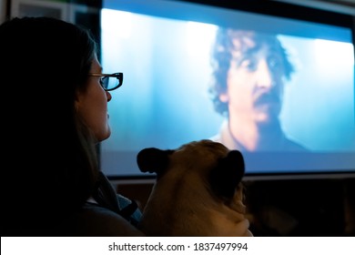 Russia Moscow 06.10.2020 Woman, Girl With Glasses With Dog Watch Tv, Movie At Night On Huge Screen At Home On Couch.Interesting Film,evening In Quarantine.Self-isolation.Covid-19 Coronavirus Pandemic.