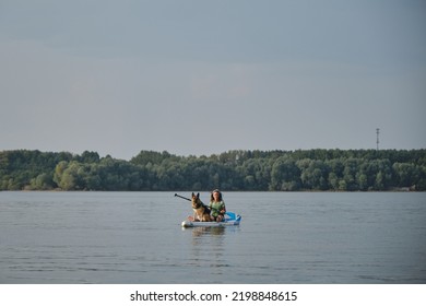 Russia, Moscow - August 2022. Young Woman Paddling In Lake Sitting On Inflatable SUP Board With German Shepherd In Life Jacket. Summer Water Activity With Pet. Stand Up Paddle Dog.