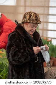 Russia, Moscow, February 2021.An Elderly Woman In A Fur Hat And Fur Coat Stands In A Flower Greenhouse With A Medical Mask And A Camera In Her Hands.