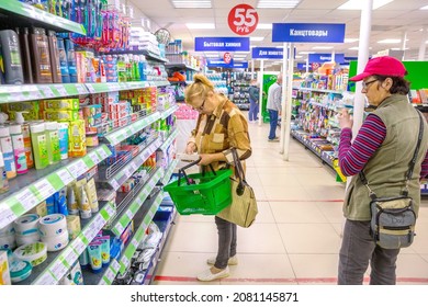 Russia Polevskoy July 2021: A Mature Woman With A Shopping Cart Stands Near A Shelf With Cleaning Products In A Store. Russian Text: Stationery, Cosmetics, Household Chemicals.