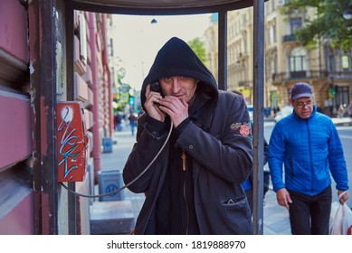 Russia, Saint Petersburg, September 2020. A Man In Black Clothes With A Hood On His Head Is Standing In A Phone Booth, Holding The Phone To His Ear And Looking At The Camera.