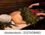 Russian bathhouse. Man in bath hat lying and sweats on wooden bench in sauna with hot steam. Hands with broom whip steamed body in spa complex. Wellness, self-care, healthy concept. Copy ad text space