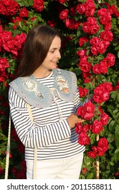 Russian Fashion Woman, Tourist Girl In Summer Garden In Suzdal Town, Russia. Look, Outfit With Embroidered Collar, Stripped Vest, White Jeans. Street Style. Red Roses, Flowers. Summer Blossom, Bloom