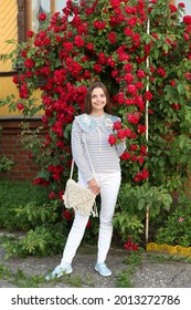 Russian Fashion Woman, Tourist Girl In Summer Garden In Suzdal Town, Russia. Look, Outfit With Embroidered Collar, Stripped Vest, White Jeans. Street Style. Red Roses, Flowers. Summer Blossom, Bloom