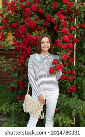 Russian Fashion Woman, Tourist Girl In Summer Garden In Suzdal Town, Russia. Look, Outfit With Embroidered Collar, Stripped Vest, White Jeans. Street Style. Red Roses, Flowers. Summer Blossom, Bloom