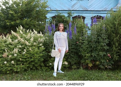 Russian Fashion Woman, Tourist Girl In Summer Garden In Suzdal Town, Russia. Look, Outfit With Collar, Stripped Vest, White Jeans. Street Style. Delphinium Flowers In Home. Suzdal Landmark, House