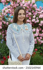 Russian Fashion Woman, Tourist Girl In Summer Garden In Suzdal Town, Russia. Look, Outfit With Embroidered Collar, Stripped Vest, White Jeans. Street Style. Pink Roses, Flowers. Summer Blossom, Bloom