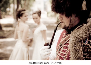 Russian hussar in vintage outfit is smoking his pipe. Two pretty women on the background are looking at him.  Retro-styled photo.
