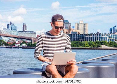 Russian man travels, works in New York, wearing striped long sleeve T shirt, cap worn backward, sunglasses, sits by river, works on laptop computer, listens music with earphone. Color filtered effect. - Powered by Shutterstock